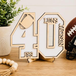 May include: A laser-cut wooden number 40 sign featuring a white background, a football graphic, and the text LIONS, JACKSON, and FCS. The sign is supported by a wooden base stand and displays a natural light wood finish with clean edges.