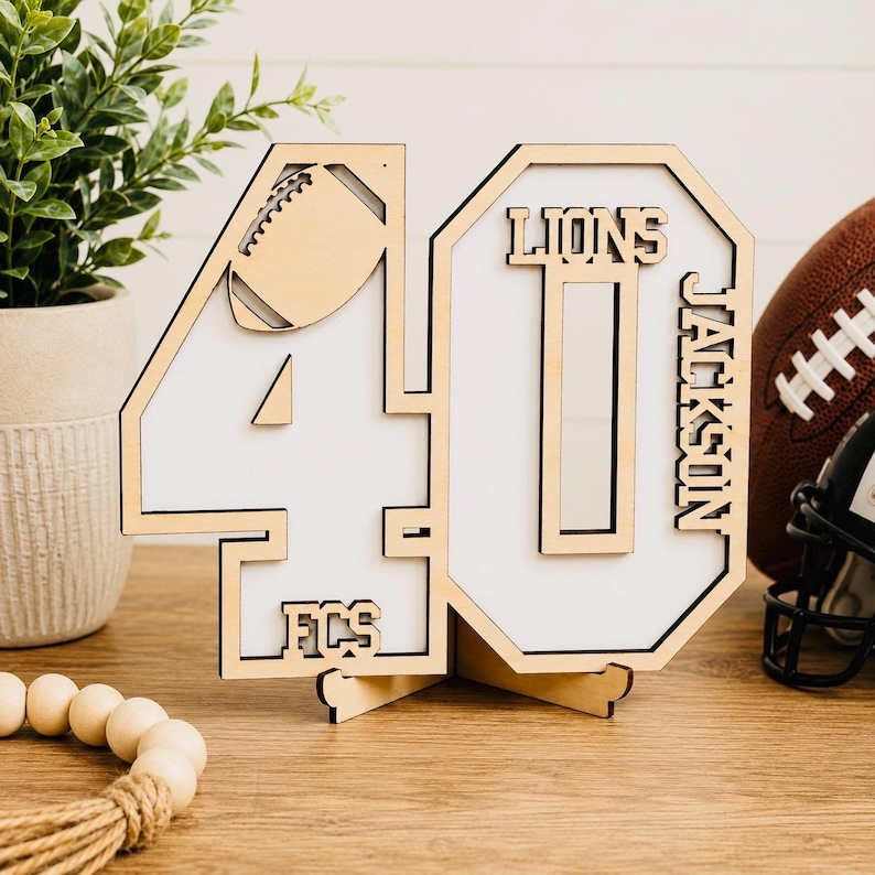 May include: A laser-cut wooden number 40 sign featuring a white background, a football graphic, and the text LIONS, JACKSON, and FCS. The sign is supported by a wooden base stand and displays a natural light wood finish with clean edges.