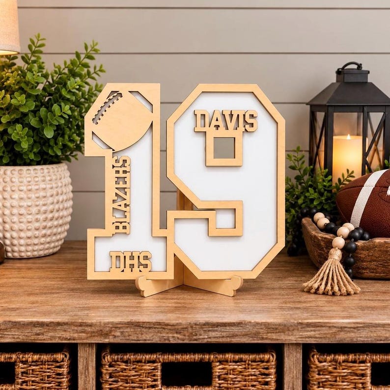 May include: A freestanding wooden number 19 sign with a white background. The number 1 features a football cutout, the text BLAZERS, and DHS. The number 9 features the name DAVIS. The sign is constructed from light-colored wood with a layered design.