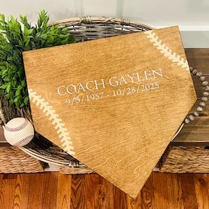 May include: Wooden home plate-shaped sign with the text "COACH GAYLEN 9/5/1957 - 10/28/2025" in white. The sign has baseball stitching details and is placed in a woven basket with a baseball and greenery.
