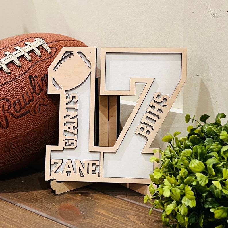 May include: A layered wooden cutout of the number 17 featuring a football icon at the top, the name ZANE, and the text GIANIS and BOHS. The design is constructed from light-colored wood with white backing panels, creating a dimensional sports-themed display.