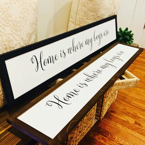 May include: Two wooden framed signs with white backgrounds and black calligraphy. The top sign reads "Home is where my boys are". The bottom sign reads "Home is where my boys are".
