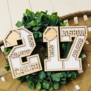 May include: Two light-colored wooden number cutouts, 2 and 17, featuring white backings. The number 2 includes the text CALEB and COMETS, while the number 17 features JOSHUA and COMETS. Both numbers are decorated with a small football icon.
