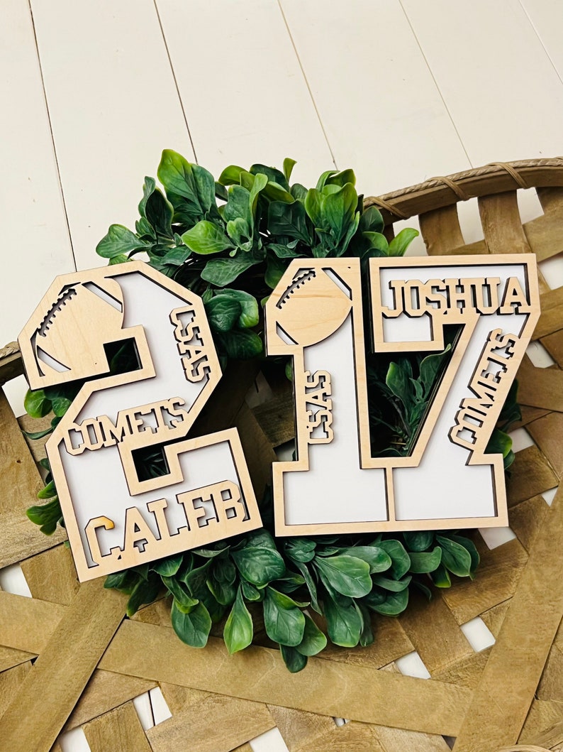 May include: Two light-colored wooden number cutouts, 2 and 17, featuring white backings. The number 2 includes the text CALEB and COMETS, while the number 17 features JOSHUA and COMETS. Both numbers are decorated with a small football icon.