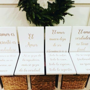 May include: Five white wooden signs with gold lettering, each with a different quote about love in Spanish. The quotes are: "El amor es paciente es bondadoso", "El Amor", "El amor nunca deja de ser", "El amor verdadero no tiene final", and "No se goza de la injusticia. más se goza de la verdad".