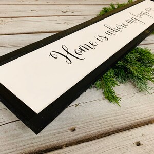 May include: A black framed wooden sign with white background and the text "Home is where my heart is" in black cursive font.