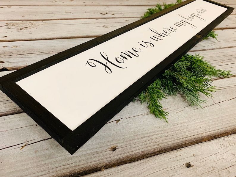 May include: A black framed wooden sign with white background and the text "Home is where my heart is" in black cursive font.
