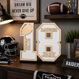 May include: A wooden number 18 display piece featuring a cutout football on the number 1, the name Seth vertically on the number 1, and the name Jake on the number 8, with a white background and light wood trim on a black base.