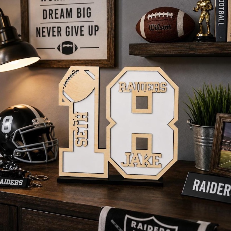 May include: A wooden number 18 display piece featuring a cutout football on the number 1, the name Seth vertically on the number 1, and the name Jake on the number 8, with a white background and light wood trim on a black base.