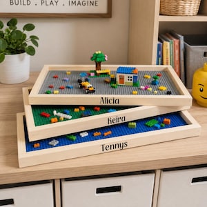 May include: Three rectangular wooden building block trays stacked vertically. Each tray features a baseplate in gray, green, or blue, with personalized names Alicia, Keira, and Tennys printed on the front edges. Small plastic bricks are scattered on top.
