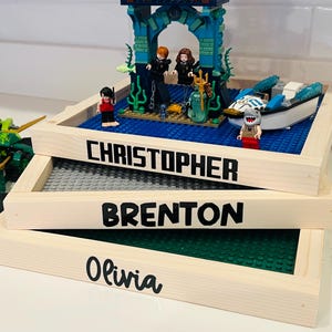 May include: Three wooden framed Lego display trays stacked on top of each other. The top tray says "CHRISTOPHER" in black letters, the middle tray says "BRENTON", and the bottom tray says "Olivia". The top tray has a Lego scene with figures and a boat.