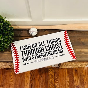 May include: White wooden sign with red stitching around the edges, resembling a baseball. The sign reads "I can do all things through Christ who strengthens me - Philippians 4:13".
