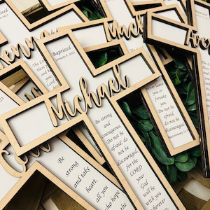 May include: Wooden cross-shaped photo frames with a white background and black frames. The frames have a cutout design with the words "Baptized" and a date, along with a name. The frames are decorated with green leaves. The frames have inspirational quotes about strength and courage.