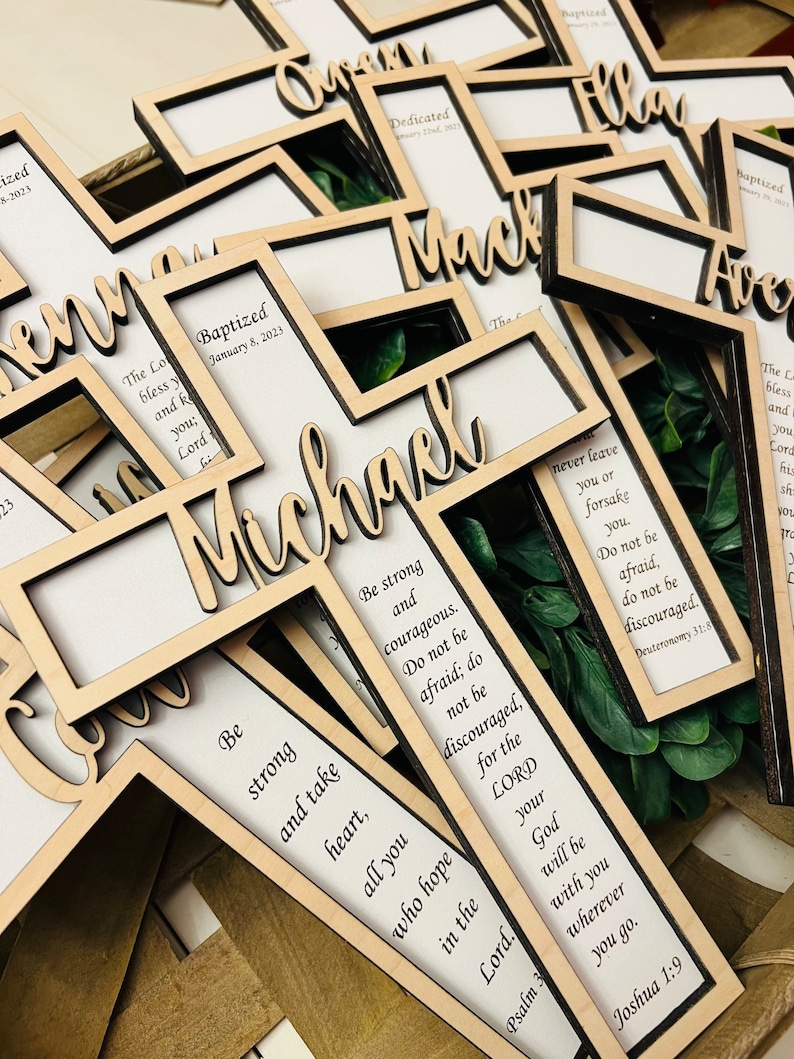 May include: Wooden cross-shaped photo frames with a white background and black frames. The frames have a cutout design with the words "Baptized" and a date, along with a name. The frames are decorated with green leaves. The frames have inspirational quotes about strength and courage.