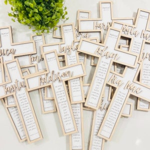 May include: A pile of wooden crosses with personalized names and a bible verse. The crosses are light brown and have a white background with black text. The crosses are all different sizes and shapes.