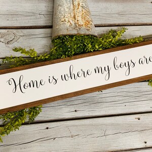 May include: A wooden sign with a white background and brown frame. The sign reads "Home is where my boys are" in black cursive lettering.