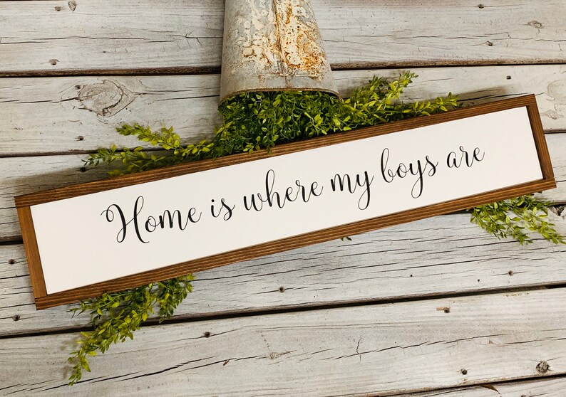 May include: A wooden sign with a white background and brown frame. The sign reads "Home is where my boys are" in black cursive lettering.