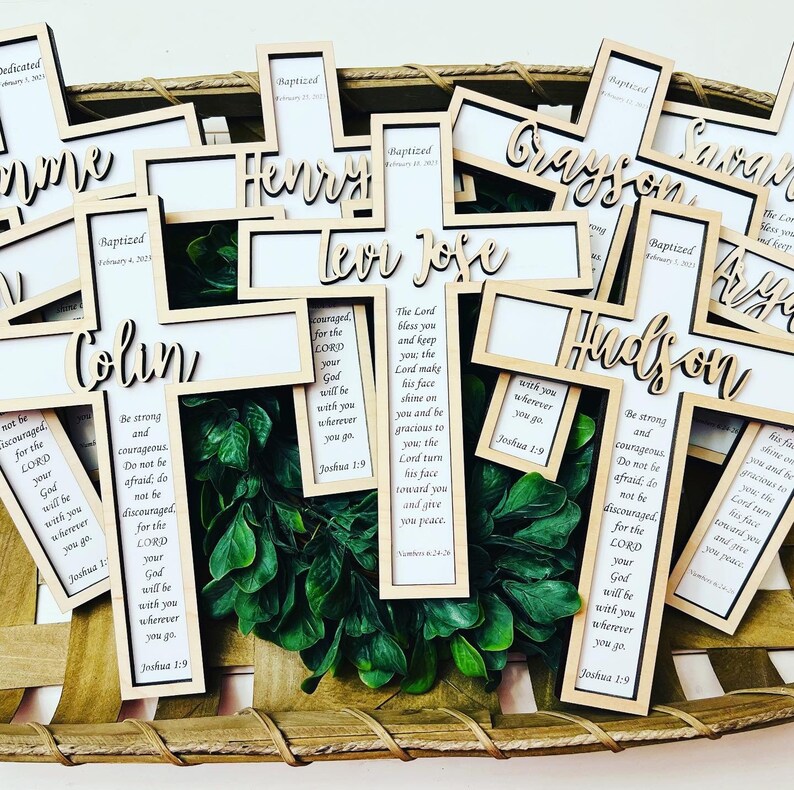 May include: A collection of wooden cross-shaped ornaments with names and bible verses. The crosses are in a woven basket with green foliage.