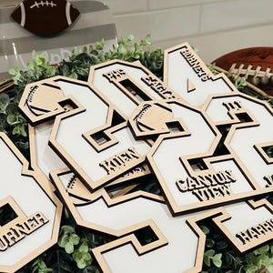 May include: A collection of laser-cut wooden jersey number ornaments featuring white inserts. Each piece displays engraved text including names like Turner, Kuen, and Harris, team names such as Hornets and Jaguars, and small football graphics.