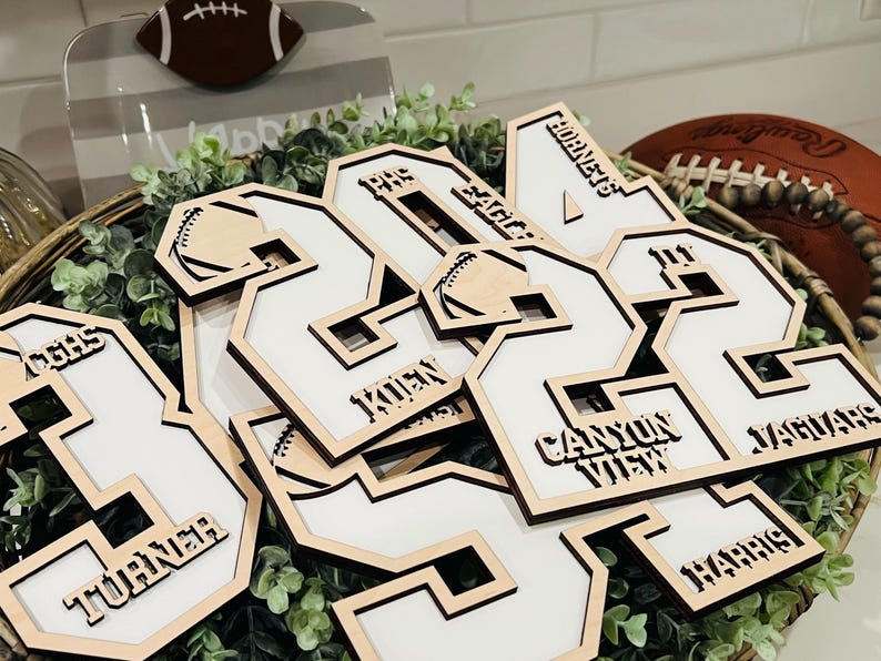 May include: A collection of laser-cut wooden jersey number ornaments featuring white inserts. Each piece displays engraved text including names like Turner, Kuen, and Harris, team names such as Hornets and Jaguars, and small football graphics.