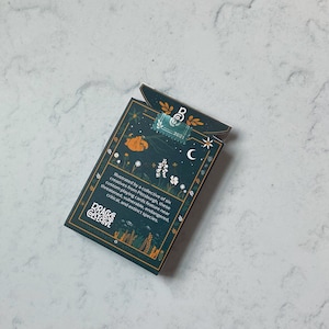 May include: A deck of playing cards with a dark teal background and botanical illustrations. The box features text that reads "DRACKS COLLECTIVE" and "2021". The design includes a sloth, flowers, and a crescent moon.