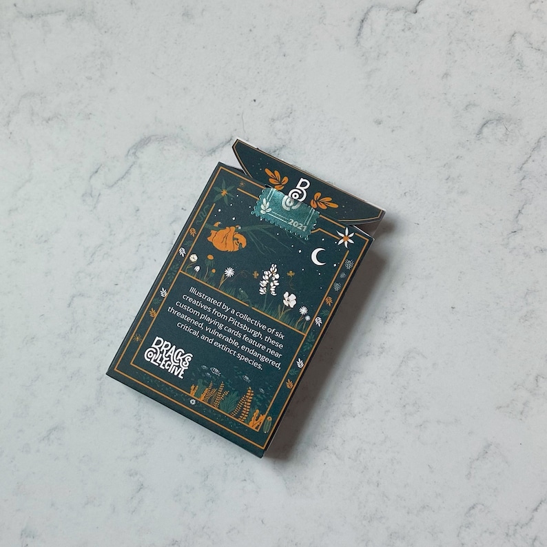 May include: A deck of playing cards with a dark teal background and botanical illustrations. The box features text that reads "DRACKS COLLECTIVE" and "2021". The design includes a sloth, flowers, and a crescent moon.
