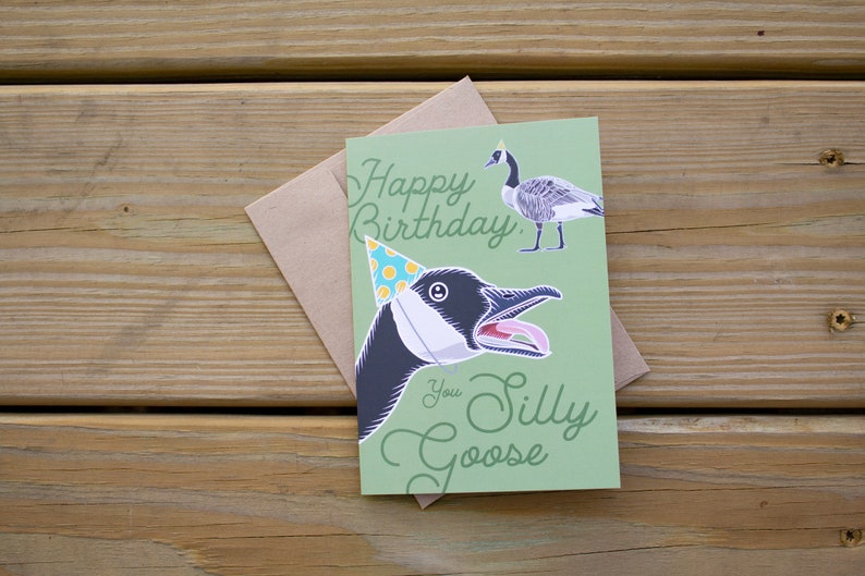 Silly goose birthday card shown head-on with kraft envelope on rustic wood surface. Green funny birthday greeting card with script text and illustrated party hat goose.