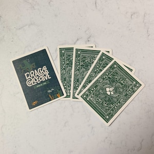 May include: A deck of playing cards with a dark teal back featuring an octopus illustration and the text "DRAGS COLLECTIVE". The card backs are green with a detailed nature-themed design. The cards are arranged on a white marble surface.