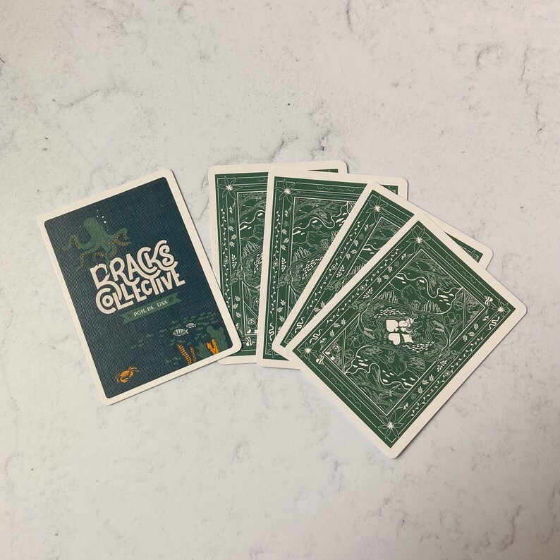 May include: A deck of playing cards with a dark teal back featuring an octopus illustration and the text "DRAGS COLLECTIVE". The card backs are green with a detailed nature-themed design. The cards are arranged on a white marble surface.