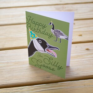 Funny goose birthday card standing upright on wood table. Green greeting card reads “Happy Birthday You Silly Goose” in script lettering and features a goose wearing a blue polka dot party hat, with a smaller full-body goose illustration in the corner.