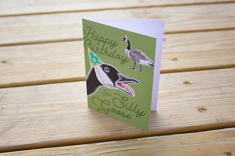 Funny goose birthday card standing upright on wood table. Green greeting card reads “Happy Birthday You Silly Goose” in script lettering and features a goose wearing a blue polka dot party hat, with a smaller full-body goose illustration in the corner.