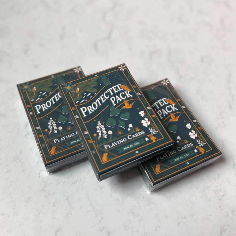May include: Three decks of playing cards with a dark teal background and a nature-themed design. The text "PROTECTED PACK" and "PLAYING CARDS" are visible. The design includes illustrations of birds, flowers, and other natural elements.