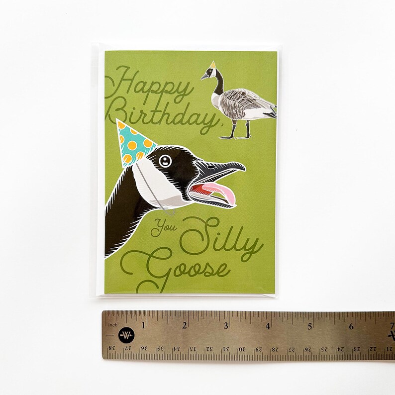 Silly goose birthday card in protective plastic sleeve displayed head-on with metal ruler showing 4.13 inch width. Green funny birthday greeting card with party hat goose illustration.
