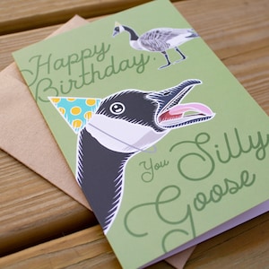 Funny goose birthday card with green background and script text that says “Happy Birthday You Silly Goose.” Features a close-up goose illustration wearing a blue polka dot party hat and a smaller full-body goose in the corner. Shown with kraft envelope on wood surface.