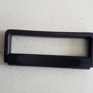 Op de afbeelding: Een zwarte kunststof handgreep, rechthoekig van vorm, met een afgeronde grip. De handgreep is ontworpen om te dragen of te bevestigen aan een tas of container. De handgreep is gemaakt van duurzaam, matzwart kunststof.