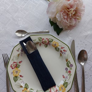 Puede incluir: Un plato de cena octogonal blanco con un patrón floral en tonos de verde, amarillo, rojo y rosa. Un porta servilletas de mezclilla azul con un tenedor y un cuchillo de plata en su interior se coloca en el plato. Una cuchara de plata y un cuchillo de plata se colocan en la mesa junto al plato.