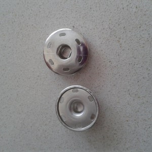 Puede incluir: Dos botones a presión de metal plateado, uno con un centro elevado y el otro con un centro empotrado.