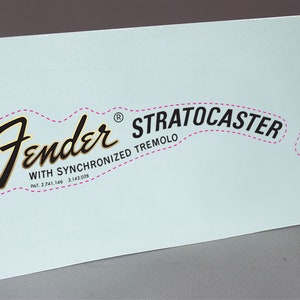 May include: A white sticker with black text that reads "Fender® Stratocaster with Synchronized Tremolo Pat. 2,741,146 3,143,078". The sticker also has a pink dashed line around the text and a small square with the text "Original Contour Body Patented" in black text.