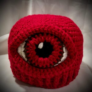 May include: Red crocheted beanie hat with a large brown eye design. The eye has a white iris and a black pupil.