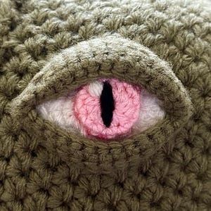 May include: Close-up of a crocheted eye with a pink iris and a black pupil. The eye is surrounded by brown crocheted fabric.