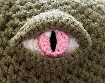 May include: A close-up of a crocheted olive green fabric featuring a textured, protruding eye design. The eye has a white sclera, a textured pink iris, and a vertical black pupil, all crafted with detailed yarn stitching and a raised eyelid.