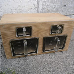 Może przedstawiać: Drewniana skrzynka z czterema przezroczystymi szklanymi pojemnikami z uchwytami. Skrzynka wykonana jest z jasnego drewna i stoi na betonowej powierzchni. Szklane pojemniki mają prostokątny kształt.