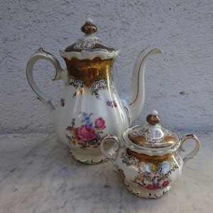 Op de afbeelding: Een vintage porseleinen koffiepot en suikerpot set. De witte stukken hebben gouden randen en bloemmotieven met roze en blauwe bloemen. De koffiepot heeft een lange, gebogen tuit en een deksel met een decoratieve knop.
