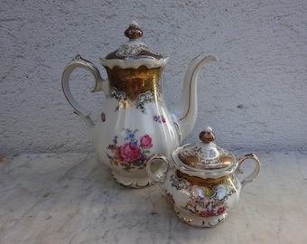 May include: A vintage porcelain coffee pot and sugar bowl set. The white pieces feature gold trim and floral designs with pink and blue flowers. The coffee pot has a tall, curved spout and a lid with a decorative finial.