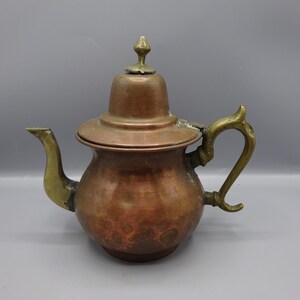 Puede incluir: Una tetera de cobre antigua con asa, boquilla y pomo de latón. La tetera tiene un cuerpo bulboso y una tapa ligeramente abovedada. El asa es ornamentada y la boquilla se curva con gracia. El cobre tiene un tono marrón rojizo cálido.