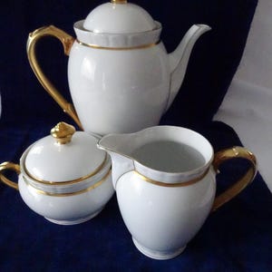 Op de afbeelding: Een witte porseleinen theeset met gouden accenten. De set bevat een theepot met deksel, een melkkan en een suikerpot met deksel. De handvatten en randen zijn goudkleurig, op een donkerblauwe achtergrond.