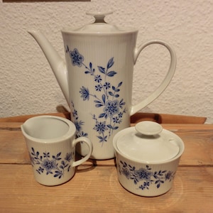 Op de afbeelding: Een witte porseleinen koffiepot, roomkan en suikerpot met een blauw bloemenpatroon. De set staat op een houten oppervlak.