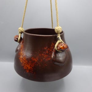Può includere: Un vaso sospeso in ceramica marrone con un motivo maculato arancione. Il vaso ha due piccole maniglie ed è sospeso da una corda beige. Il vaso è rotondo con un'ampia apertura, adatto alle piante.