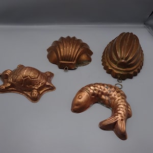 Op de afbeelding: Vier vintage koperen mallen in de vorm van een schildpad, een schelp, een sint-jakobsschelp en een vis. Elke mal heeft een lus om op te hangen. De mallen hebben een warme koperkleur en lijken gebruikt.