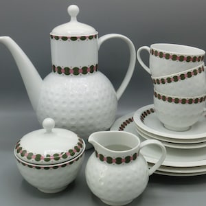 Op de afbeelding: Een wit theeservies met een textuur, een gekarteld ontwerp. De set bevat een theepot, een roomkan, een suikerpot en vier kopjes en schoteltjes. De kopjes en schoteltjes hebben een groen en roze geometrisch patroon.